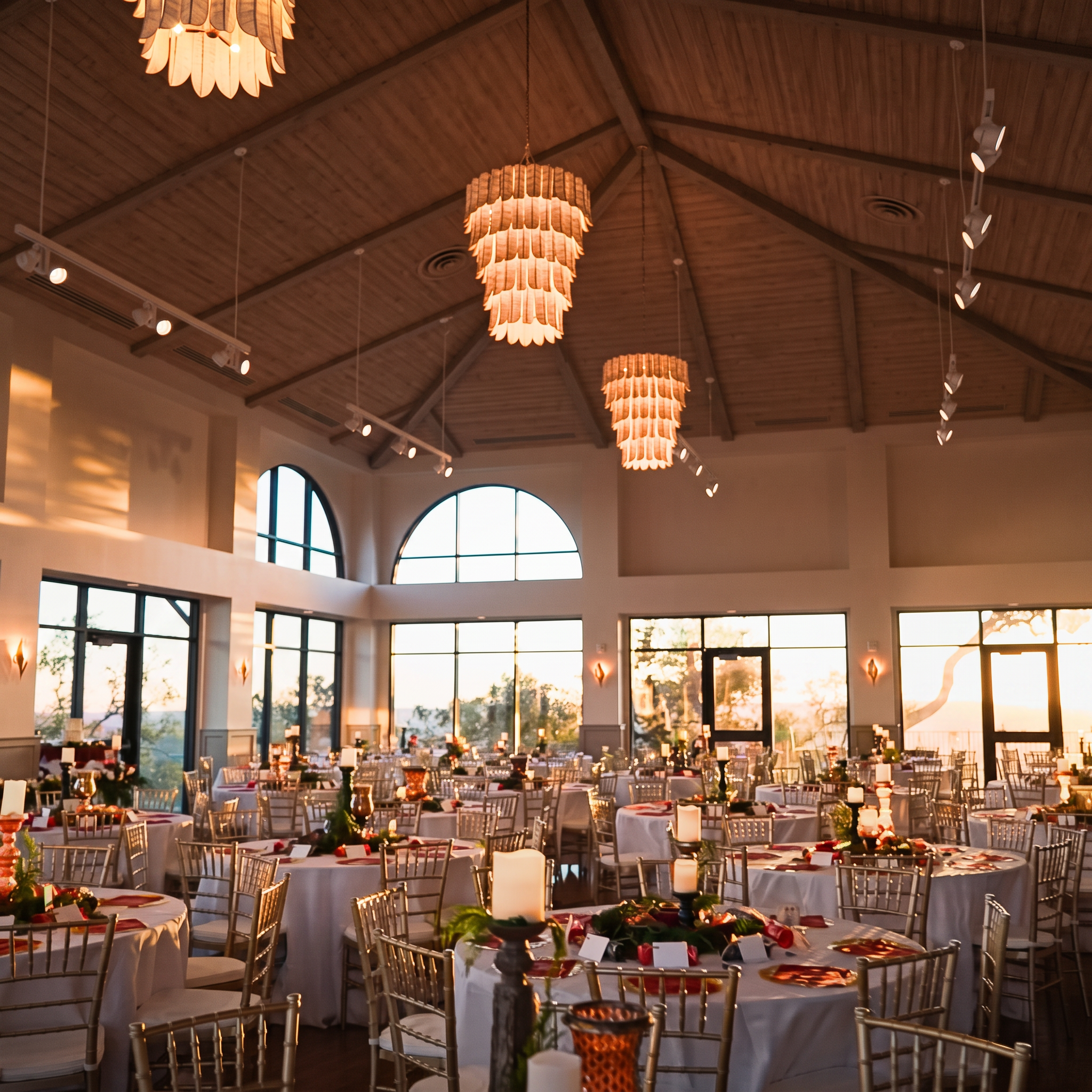 The ballroom at The Pinnacle at Westlake glowing at sunset — chandeliers overhead, tables set, and Hill Country light through arched windows.